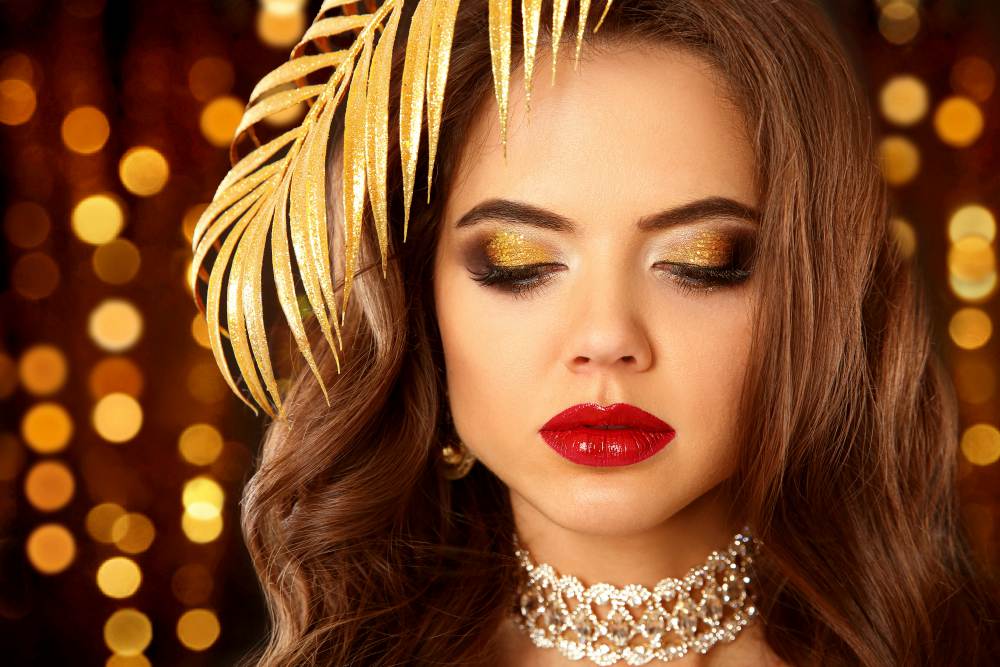 Sexy elegant woman with red lips, glitter eyeshadow in expensive diamond necklace over Christmas lights background | New Year’s Eve Makeup Tutorial | Gold Glitter Cut Crease With Red Lips 