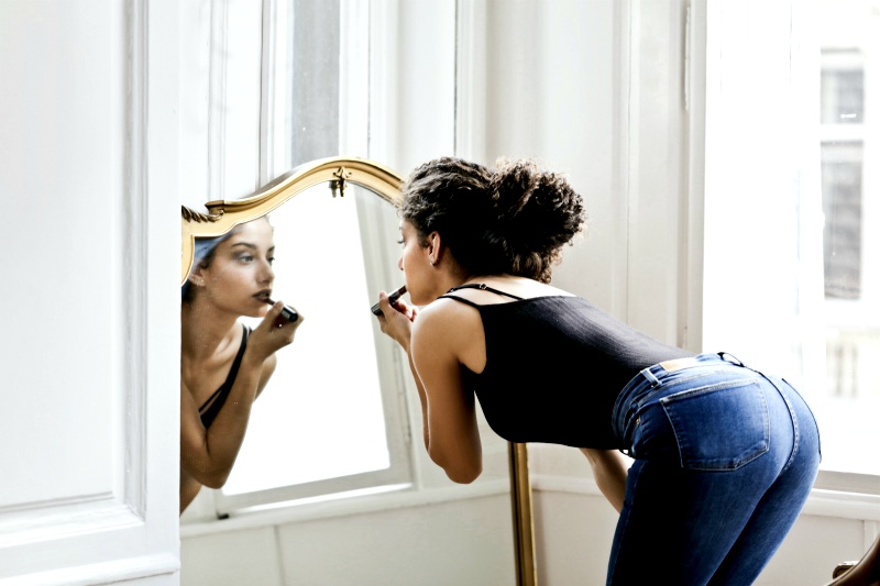 photo of woman in black tank top and blue denim applying lipstick in front of mirror | fall makeup looks