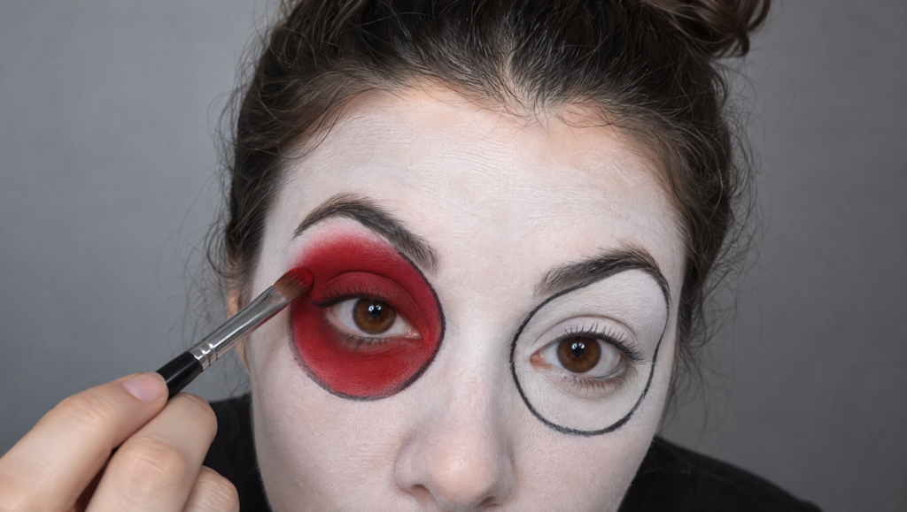 Day of the Dead makeup tutorial step 5: flat brush filling one outlined eye socket with deep red color